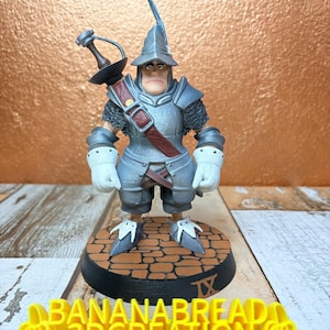 May include: A 3D-printed figurine of a medieval knight in silver armor, a red strap, and a sword. The figurine stands on a circular base with a brick pattern. The base has the Roman numeral IX. The text "BANANABREAD 3DCREATION" is in yellow.