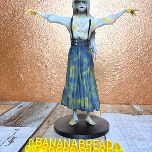 May include: A 3D-printed figurine of a woman with outstretched arms, wearing a white blouse, blue skirt, and black suspenders. The figure has pale skin and long white hair. The base is black, and the text "BANANABREAD 3DCREATION" is visible.