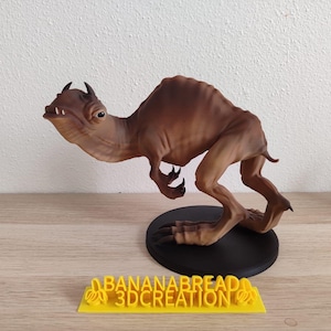 Puede incluir: Una figurita de color marrón, detallada, impresa en 3D de una criatura con cuello largo y cabeza puntiaguda. La figurita está en posición de carrera y está de pie sobre una base negra. La base tiene el texto "BANANABREAD 3DCREATION" impreso en amarillo.