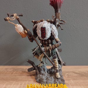 May include: A detailed figurine of a warrior with a white fur-like collar and a red emblem. The figure holds a staff and a weapon, with a red plume on its back. The base reads "BANANABREAD 3DCREATION".
