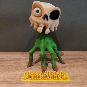 May include: A 3D printed figurine of a green, skeletal hand with a single, large eye and a black patch over the other eye. The figurine is standing on a yellow base with the text "BANANABREAD 3DCREATION" printed on it.