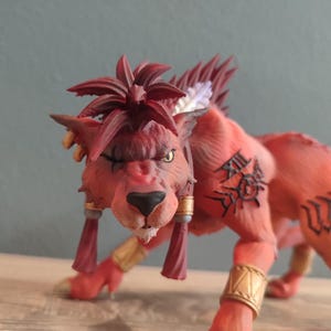 May include: A red and orange figurine of a lion-like creature with a fierce expression. The figurine has detailed features, including a spiky mane, tribal tattoos, and gold accents. The creature is in a crouching position.