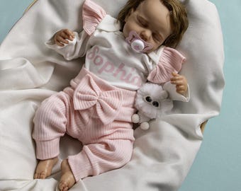 May include: A pink and white baby outfit with a bow on the pants and the name "Sophia" on the shirt. The baby is sleeping in a white basket.