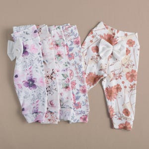 May include: Five pairs of floral print leggings for babies or toddlers. The leggings are white with pink, purple, and orange floral patterns. One pair of leggings has a white bow on the waistband.