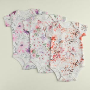 May include: Three white baby onesies with floral prints. The onesies have short sleeves and snap closures. The floral prints are in shades of pink, purple, and orange.