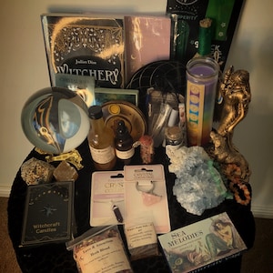 May include: A selection of spiritual items, including books, crystals, candles, and herbs, displayed on a black surface. Items include a crystal ball, a gold-coloured statue, and a book titled "Witchery".