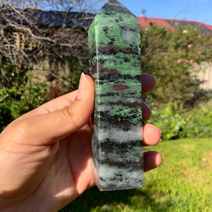 May include: A polished crystal tower featuring a mix of vibrant green, deep red, and black mineral inclusions with translucent white quartz bands. The stone has a hexagonal prism shape with a pointed top, displaying natural colour variations throughout.