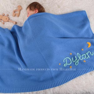 May include: A blue knitted baby blanket with a crescent moon and stars embroidered on it. The blanket has the name "Dylan" embroidered in green on it. The text "Handmade Products from HulkeBar" is also embroidered on the blanket.
