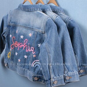 May include: Three light blue denim jackets hanging on wooden hangers. Each jacket features embroidered details, including the name "Sophia" in pink script, flowers, a sun, and a rainbow. Classic denim jackets.
