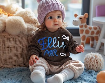 May include: A baby wearing a brown sweater with the name "Ellie" in blue script, paired with beige pants and a lavender knit hat with a pom-pom. The baby is sitting on a blue rug, with a giraffe toy and a basket in the background.