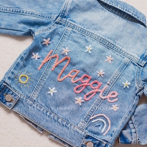 Op de afbeelding: Lichtblauwe spijkerjas met borduursels. De achterkant heeft de naam "Maggie" in roze script, omringd door geborduurde bloemen, een zon en een regenboog. Een stijlvolle en gepersonaliseerde kinderjas.