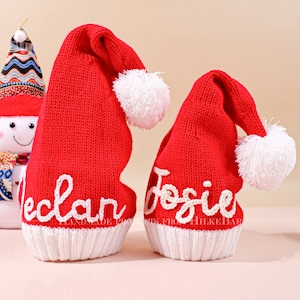 Può includere: Due cappelli di Babbo Natale in maglia rossa con pon pon bianchi e bordino bianco. I cappelli sono personalizzati con i nomi "Declan" e "Josie" in ricamo bianco.