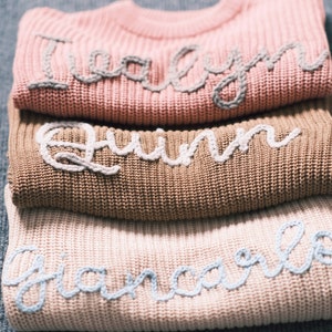 May include: Three knitted jumpers stacked on top of each other. The top jumper is pink with the name "Talyn" embroidered in white. The middle jumper is brown with the name "Quinn" embroidered in white. The bottom jumper is light pink with the name "Giancarlo" embroidered in light blue.