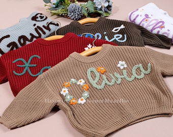 May include: A collection of children's sweaters in various colors, including brown, red, and light blue. Each sweater features a name embroidered on the front in a contrasting color, with floral or other decorative accents. The sweaters are displayed on wooden hangers.