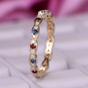 Può includere: Un anello dorato con un motivo ripetuto di pietre preziose rosse, blu e bianche. L'anello presenta una serie di disegni ovali, ognuno contenente una piccola pietra preziosa. Le pietre preziose sono incastonate in una fascia dorata.