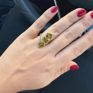 Könnte beinhalten: Ein goldener Ring mit drei ovalen, olivgrünen Edelsteinen. Das Design des Rings ist eine moderne Interpretation eines klassischen Stils, bei der die Steine linear angeordnet sind. Der Ring wird an einer Hand mit rotem Nagellack gezeigt.