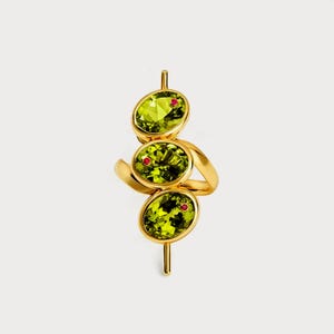 May include: A gold ring featuring three oval-cut, light green gemstones with small red accents. The ring's design is a unique, modern style, with the gemstones arranged vertically and connected by a gold band.