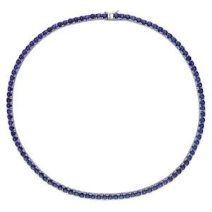 Puede incluir: Un collar de tenis con una hilera continua de piedras preciosas redondas de color azul oscuro. Las gemas están engarzadas juntas en un metal plateado, creando un círculo completo. El collar se asegura con un cierre en la parte posterior.