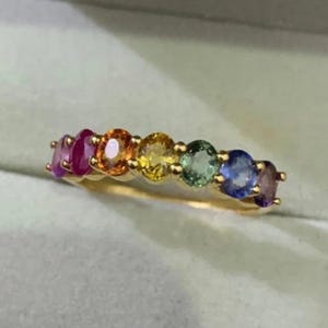 Könnte beinhalten: Ein goldener Ring mit einer Reihe oval geschliffener Edelsteine in Regenbogenfarben: Lila, Rot, Orange, Gelb, Grün, Blau und Lila. Die Edelsteine sind in einem goldenen Band gefasst.