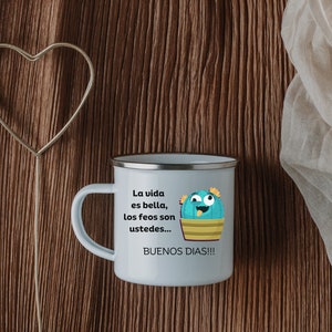 May include: A white enamel mug with a silver rim and handle. The mug features a cartoon cactus with the text "La vida es bella, los feos son ustedes... BUENOS DIAS!!!" in black. The mug is set against a brown wooden background.