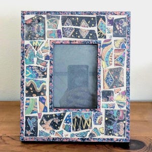 May include: A rectangular photo frame with a mosaic design. The frame is composed of various colorful, irregularly shaped pieces, including blue, green, and yellow. The frame's border is a mix of pink and blue. The frame is sitting on a wooden surface.