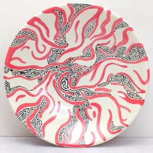 May include: A round, off-white ceramic plate with a vibrant, abstract design. The pattern features flowing, wavy lines in bright red, intertwined with black, intricate shapes. The plate's rim is smooth, and the overall aesthetic is modern and artistic.