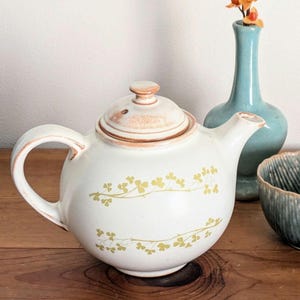 May include: White ceramic teapot with a rounded body, a curved handle, and a lid. The teapot is decorated with a yellow floral pattern. A small blue vase with orange flowers and a blue bowl are also in the image.