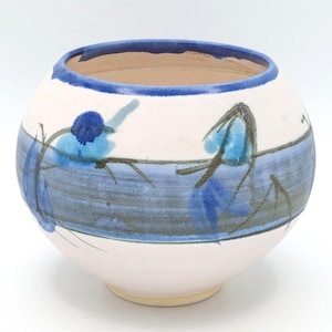 May include: A round, handmade ceramic bowl with a wide opening. The bowl is primarily white with a horizontal blue band and blue floral designs. The rim is dark blue, and the interior is a light brown color.