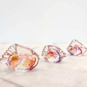 May include: Three clear glass fish figurines of varying sizes, each featuring internal swirls of orange, red, and purple color. The glass bodies are translucent with defined fin details, arranged in a row on a light-colored surface.