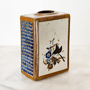 May include: A rectangular ceramic vase with a speckled off-white front panel featuring a hand-painted blue and brown floral motif. The side panel displays a dense grid of small blue dots. The edges and top are finished in a glossy brown glaze.