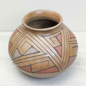 May include: A round, tan-colored pottery vessel with a wide opening. The exterior features a geometric design in black, red, and light tan. The design consists of triangles and lines. The pottery has a smooth surface and a rustic appearance.