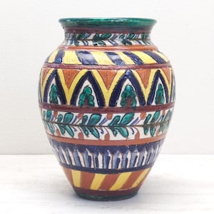 May include: A decorative ceramic vase with a wide body and a narrow neck. The vase features a colorful design with yellow, blue, orange, and green geometric patterns and leaf motifs. The rim is green.