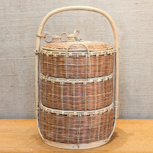 May include: A three-tiered woven wicker basket with a rounded handle. The basket has a natural brown color and a lid. The handle is attached to the sides of the basket with a decorative chain. The basket is sitting on a wooden surface.