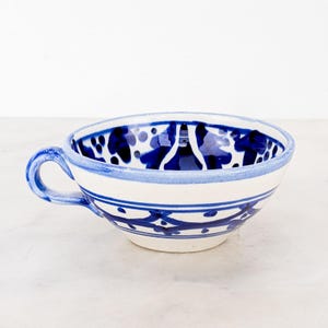 May include: A ceramic bowl with a single handle, featuring a white base color and hand-painted blue decorative patterns. The exterior and interior display blue horizontal lines, floral motifs, and dotted accents around the rim and body.