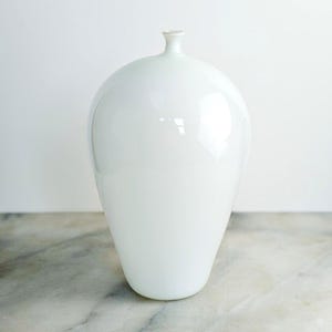 May include: A smooth, opaque white glass vase with a rounded, bulbous body that tapers toward a narrow, short neck and a small, circular opening at the top. The surface of the vase has a glossy finish that reflects light, creating subtle highlights.