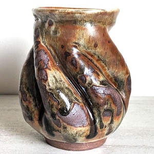 May include: A handcrafted ceramic vase with a unique, twisted design. The vase features a blend of earthy tones, including brown, green, and black, with a glossy finish. The vase has a small, round base and a slightly flared top, perfect for displaying flowers or as a decorative piece.