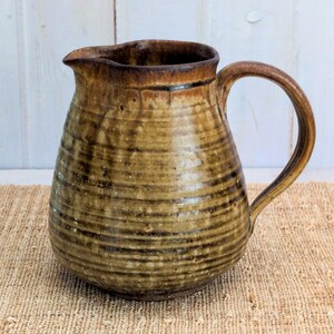 May include: A handcrafted ceramic pitcher with a wide base and a curved handle. The pitcher has a brown and tan color scheme with horizontal textured lines. The spout is slightly angled for easy pouring. The pitcher is likely used for serving beverages.