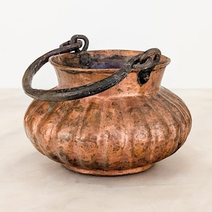 May include: A round copper pot with a fluted, ribbed body and a dark, weathered patina. The vessel features a wide, open mouth and a thick, curved dark metal handle attached to two circular loops on opposite sides of the rim.