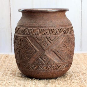 May include: A brown, textured ceramic pot with a wide, rounded body and a smooth, dark rim. The exterior surface features intricate, carved geometric patterns, including repeating diamond shapes, linear hatch marks, and stylized floral motifs.