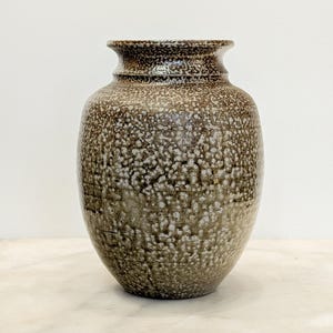 May include: A ceramic vase with a rounded body and a flared rim, featuring a mottled glaze in shades of brown, gray, and white. The surface has a textured, speckled appearance with subtle horizontal lines visible across the exterior of the vessel.