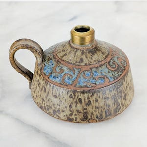 May include: A round ceramic oil lamp with a speckled tan and brown glaze. It features a single curved handle on the side and a decorative band of blue and brown swirling patterns around the top. A brass collar is centered at the top opening.