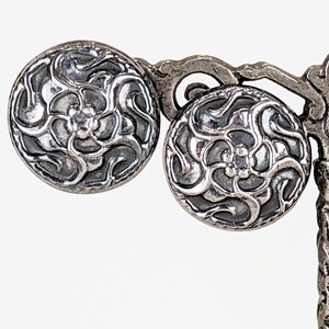 May include: A pair of round silver-toned metal buttons featuring a raised, swirling floral pattern with dark recessed areas. The circular buttons have a polished metallic finish and a detailed, intricate design that radiates from the center.