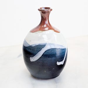 May include: A ceramic vase with a rounded body and a narrow neck. The top section is a glossy reddish-brown, transitioning into a cream-colored middle. The lower portion features a deep navy blue glaze with white drip-like patterns across the surface.