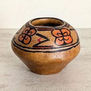 May include: A small, hand-painted ceramic pot with a rounded body and a wide opening. The pot is tan with black, red, and orange floral and geometric designs. The rim is dark brown. The pot is likely used for decorative purposes.