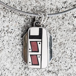 May include: A silver pendant necklace with a rectangular pendant. The pendant features a geometric design with black, white, and maroon shapes. The necklace has a silver chain.