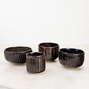 May include: Four ceramic bowls and cups with a dark, glossy, metallic-toned glaze. The vessels feature vertical ribbed textures along the exterior walls, with a smooth, light-colored base visible on the bottom of each individual piece.
