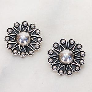 May include: A pair of silver-toned stud earrings. Each earring features a central, rounded design surrounded by a floral pattern of small, raised spheres and pointed petals. The earrings are set against a light background.
