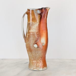 May include: A tall ceramic pitcher with a curved handle and a flared spout. The surface features a mottled glaze in shades of burnt orange, cream, and earthy brown, with small, raised shell-like textures embedded into the side of the vessel.
