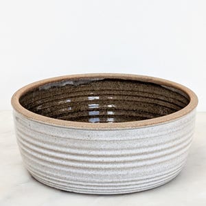 May include: A round ceramic bowl featuring a textured exterior with horizontal ridges and a speckled off-white glaze. The interior of the bowl is finished with a smooth, dark brown glossy glaze, and the rim shows the natural tan clay color.