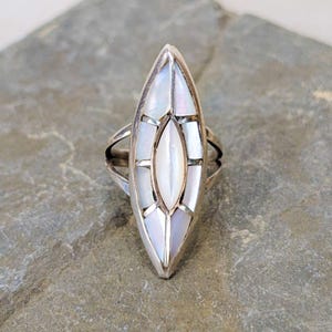 May include: A silver ring featuring a long, marquise-shaped centerpiece inlaid with multiple segments of iridescent white mother-of-pearl. The metal frame surrounds each individual shell piece, creating a geometric, elongated design on a split band.